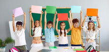 Happy Elementary School Boys And Girls Hold Up Colored Paper Sheets In Classroom. Cheerful Preteen Schoolchildren In Casual Clothes Stand In Row With Raised Speech Bubbles With Copy Space. Web Banner.
