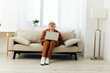 © Tatiana - a cheerful elderly woman with a short haircut, a businesswoman is sitting on a cozy sofa near the window in a brown suit and holding a video conference on a laptop. Photo in full