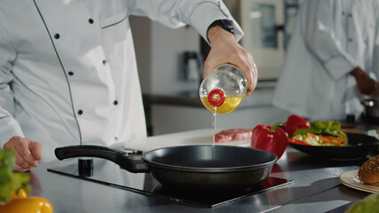 Wall Mural - Male chef pouring sunflower oil from bottle in pan on stove, preparing frying liquid to make delicious gourmet meal. Professional man cooking meat and organic vegetable recipe. Close up.