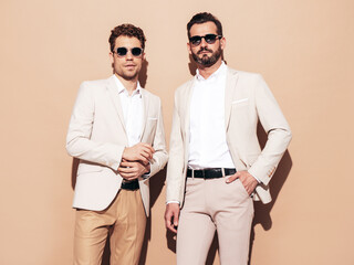 Portrait of two handsome confident stylish hipster lambersexual models. Sexy modern men dressed in white elegant suit. Fashion male posing in studio near beige wall in sunglasses