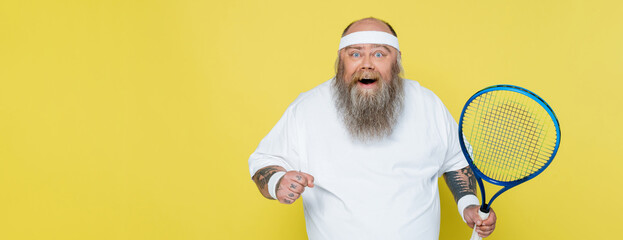 Wall Mural - excited overweight man with tennis racquet looking at camera isolated on yellow, banner.