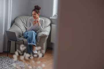 Wall Mural - smiled beautiful young mixed-race woman sitting at home with cup of tea at laptop.