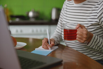Wall Mural - female holding cup of tea in hand and writing in notebook while sitting at table