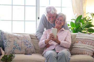 Smiling beautiful caucasian senior couple sitting at home using smart phone, old people and new technologies concept