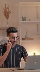 Vertical video. Late work. Busy man. Mobile agreement. Casual smiling guy talking smartphone sitting desk looking opened laptop light room interior.