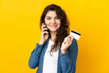 Wall Mural - Teenager Russian girl isolated on yellow background keeping a conversation with the mobile phone and holding a credit card