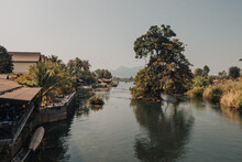 Giant Tree Coming Out Of The Water On The Mekong River, Don Det, Laos On A Sunny Day With Houses On The Banks Of The River. 