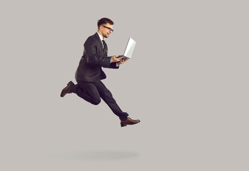 Successful joyful young businessman running with laptop in hurry to boost his career. Excited man in suit and glasses runs and jumps with laptop on gray background. Full length. Banner.