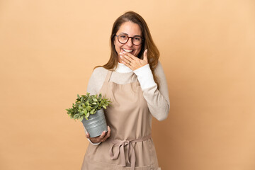 Wall Mural - Middle age gardener woman holding a plant isolated on beige background happy and smiling covering mouth with hand