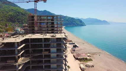 Wall Mural - Drone view of construction site, multi-storey buildings under construction against backdrop of sea and mountains on sunny summer day. Construction equipment, crane and workers walk around building.