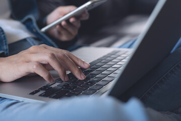 Canvas Print - Young casual woman, freelancer in blue jeans and sweater sitting on sofa using mobile phone, working on laptop computer at home, close up