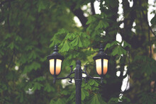 Old Cast Iron Lamp Post With Two Warm Lanterns On Trees Background In Evening. Concept Of Nostalgia By Romantic Meeting In Park