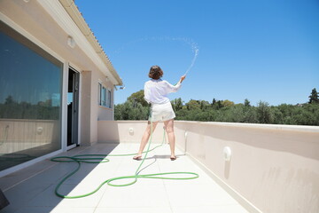 A mature woman in white clothes clean refresh a terrace of villa with water hose