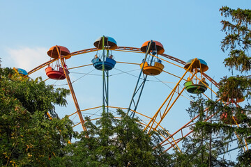 Old ferris wheel. Multi-colored attraction in the trees. Amusement park. Ferris wheel against the blue sky. Metal carousel design