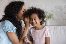 Cheerful African Girl Listens Mothers Secret Sit Together On Couch, Enjoy Trustworthy Conversation, Having Friendly Communication. Young Mom Whispering In Ear, Share Gossips To Cute Smiling Daughter