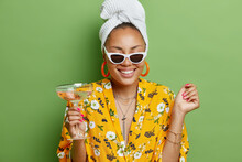 Positive Young Female Model With Manicure Smiles Toothily Wears Sunglasses White Towel Wrapped On Head And Dress Enjoys Drinking Cocktail Has Happy Mood Isolated Over Bright Green Background.