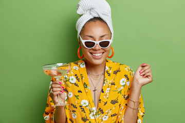 Wall Mural - Positive young female model with manicure smiles toothily wears sunglasses white towel wrapped on head and dress enjoys drinking cocktail has happy mood isolated over bright green background.