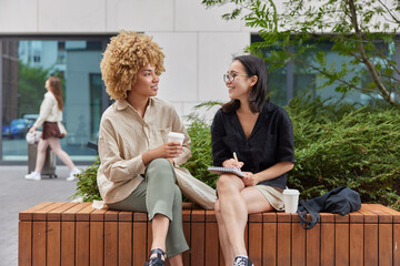 Happy Asian female journalist meets with famous person to take interview writes down notes in notebook drink takeaway coffee have pleasant conversation pose together against urban background