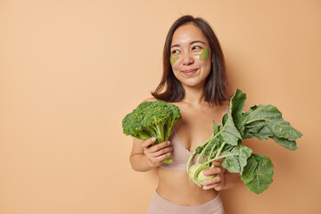 Beautiful Asian woman keeps to healthy nutrition holds green broccoli and kohlrabi going to make fresh vegetable salad applies hydrogel patches under eyes wears top leggings isolated over beige wall