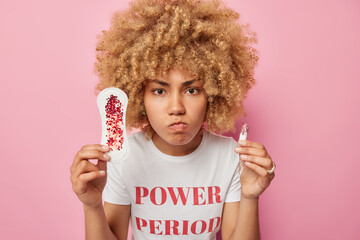 Wall Mural - Horizontal shot of displeased curly woman has menstruation chooses hygiene product holds sanitary napkin and tampon selects best absorbency item dressed casually isolated over pink background