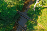 Fototapeta  - Nizinna mała rzeka. Jest słoneczny dzień. Brzegi porośnięte trawą. Nad rzeką przerzucona jest kładka dla pieszych. Jest słoneczny dzień. Zdjęcie z drona.