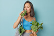 Leinwandbild Motiv Dark haired slim Asian woman keeps to healthy diet drinks homemade fresh green smoothie carries vegetables in net bag isolated over blue background. Vegan meal super foods and detox concept.