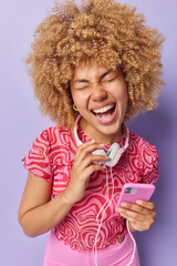 Wall Mural - Overjoyed pretty teenage girl with curly bushy hair wears stereo headphones around neck uses phone listens favorite audio track cannot imagine her life without music has upbeat mood poses indoors