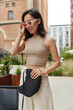 © WHstudio Leushin N - Stylish brunette Asian woman wears sunglasses t shirt and trousers holds knitted bag focused down stands outdoors in urban settings strolls during sunny summer day. Street lifestyle concept.