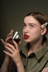 Wall Mural - Portrait of a young attractive girl with a smooth hairstyle and clips on her brown hair in a green stylish dress with a glass filled with brown liquid in her hands. Close-up, green background