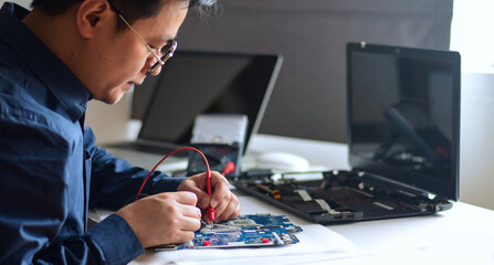 young man who is a computer technician A laptop motherboard repairman is using an IC meter to look for defects on the motherboard to repair on his desk. Board repair with modern technology