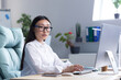 © Liubomir - Portrait of a young beautiful Asian woman in the office. Businesswoman, director, freelancer in a white shirt and glasses sits at a desk, works at a computer. Serious, looking at the camera.