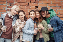 Several Happy Intercultural Teenagers With Glasses Of Soda Standing Close To One Another Against Brick Wall And Looking At Camera