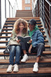 © Seventyfour - Smiling teenage couple in casualwear looking through lecture notes in copybook while sitting on long staircase by modern building