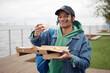 © Seventyfour - Happy black guy in casualwear holding slice of pizza over square box while having snack and looking at camera during picnic in park