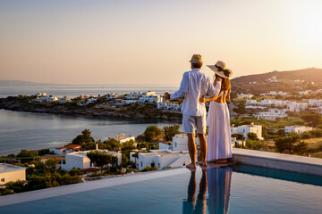 Happy couple on vacation time enjoys the summer sunset over the Aegean Sea by the swimming pool with an aperitif drink