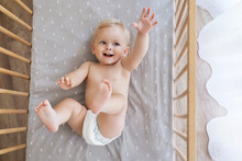 Upper View Of Little Playful Baby Boy In Diaper Lying On His Back Reaching Hand Out To Touch Toy Or Advertising Text, Playing In His Bed. Childcare Concept. Happy Childhood
