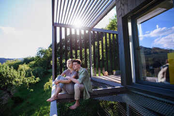 Young couple sitting and cuddling in hammock terrace in their new home in tiny house in woods, sustainable living concept.