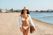 © Syda Productions - people, summer holidays and leisure concept - happy young woman in bikini swimsuit, white shirt and straw hat with bag walking along beach