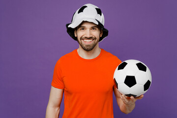 Wall Mural - Young smiling cheerful happy cool fun fan man he wears orange t-shirt cheer up support football sport team hold in hand soccer ball panama hat watch tv live stream isolated on plain purple background.