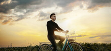 Young Man Rides A Bicycle Across A Field In A Village At Sunset. Romantic Retro Bike Ride