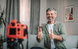 © Prostock-studio - Glad happy caucasian old male blogger with beard showing thumbs up and shooting video in living room