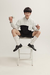 Wall Mural - Portrait of stylish young man with piercing sitting on chair, drinking coffee isolated over grey studio background