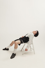 Wall Mural - Portrait of young man in black and white outfit sitting on chair, posing isolated over grey studio background. Looks relaxed