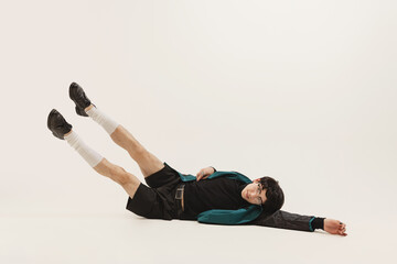 Wall Mural - Portrait of stylish young man in black outfit and green coat posing, lying on floor isolated over grey studio background