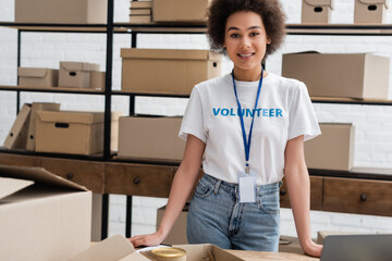 Sticker - young african american volunteer with name tag smiling at camera near carton boxes.