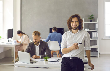 Wall Mural - Portrait of happy bearded young business man with long wavy hair holding pen, leaning on office desk, looking at camera and smiling with employees working in background