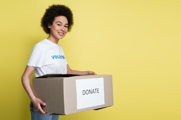 Wall Mural - smiling african american volunteer carrying donate box isolated on yellow.