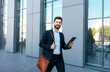 © Prostock-studio - Smiling attractive european millennial bearded businessman in suit walks with bag and tablet on street