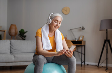 Wall Mural - Senior woman using smartphone and wearing headphones, sitting on fitball during online domestic workout