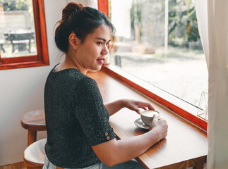 Beautiful Asian woman sits down at the bar counter inside a window coffee shop holding a cup of coffee, smiling relaxed in a cafe